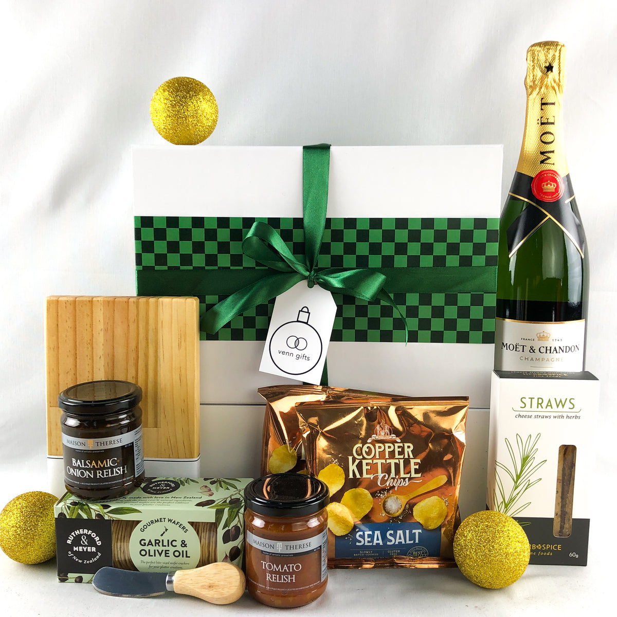 A curated gift box arrangement on a white background featuring a white gift box with a green checkered band and ribbon, a bottle of Moët & Chandon Champagne, a bag of Copper Kettle Sea Salt, jars of relish, a wooden cutting board by Venn Gifts.