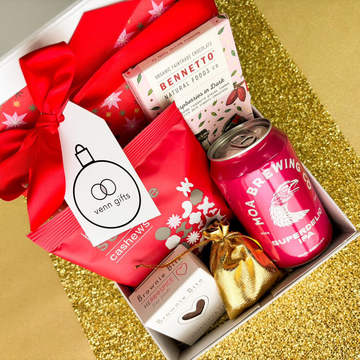 An assortment of chocolate, snacks and Moa Brewing IPA beer on a white giftbox with red ribbon against a gold background by Venn Gifts.