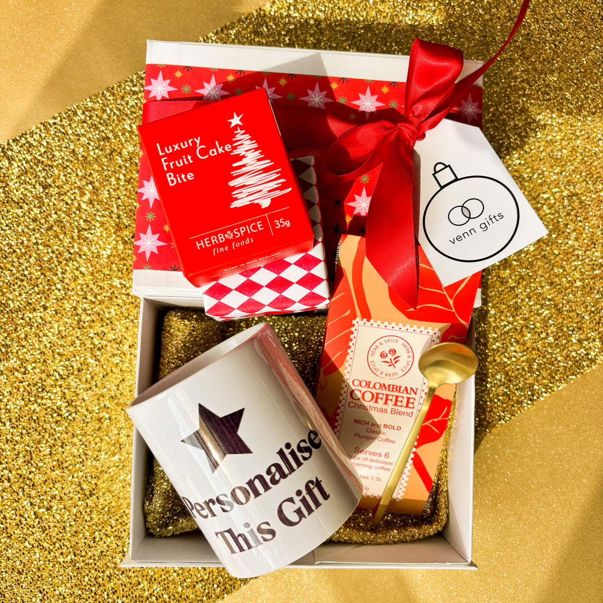 White gift box with red wrapping and red bow with coffee, gold teaspoon and personalised mug and gift wrapped christmas fruit cake against a gold background by Venn Gifts.
