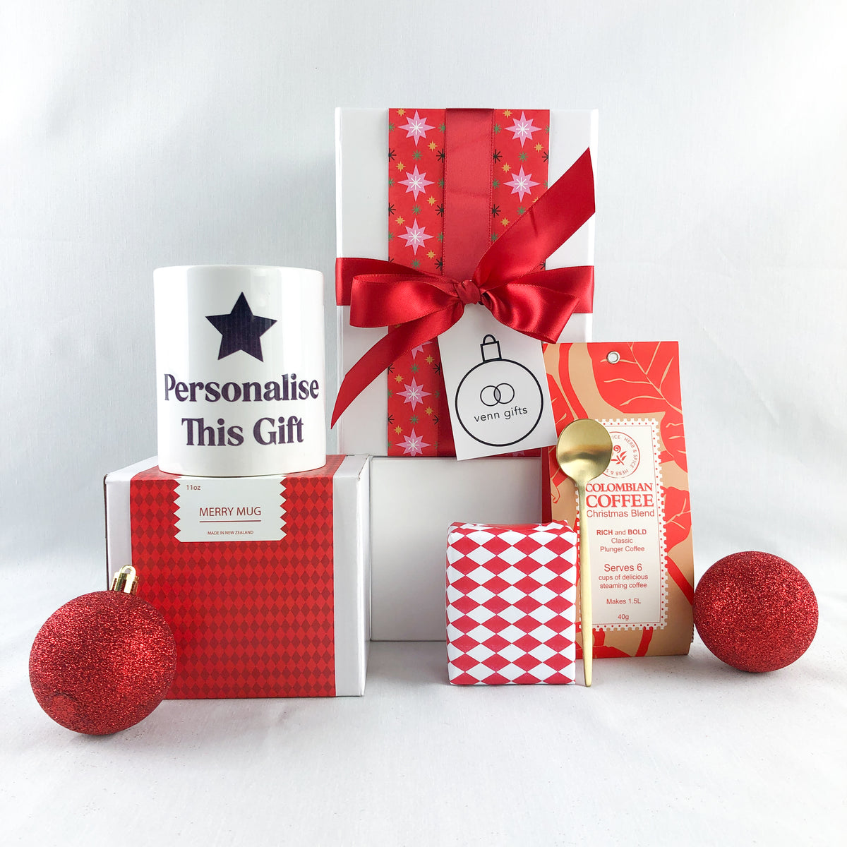 White gift box with red wrapping and red bow with coffee, gold teaspoon and personalised mug and gift wrapped christmas fruit cake against a white background by Venn Gifts.