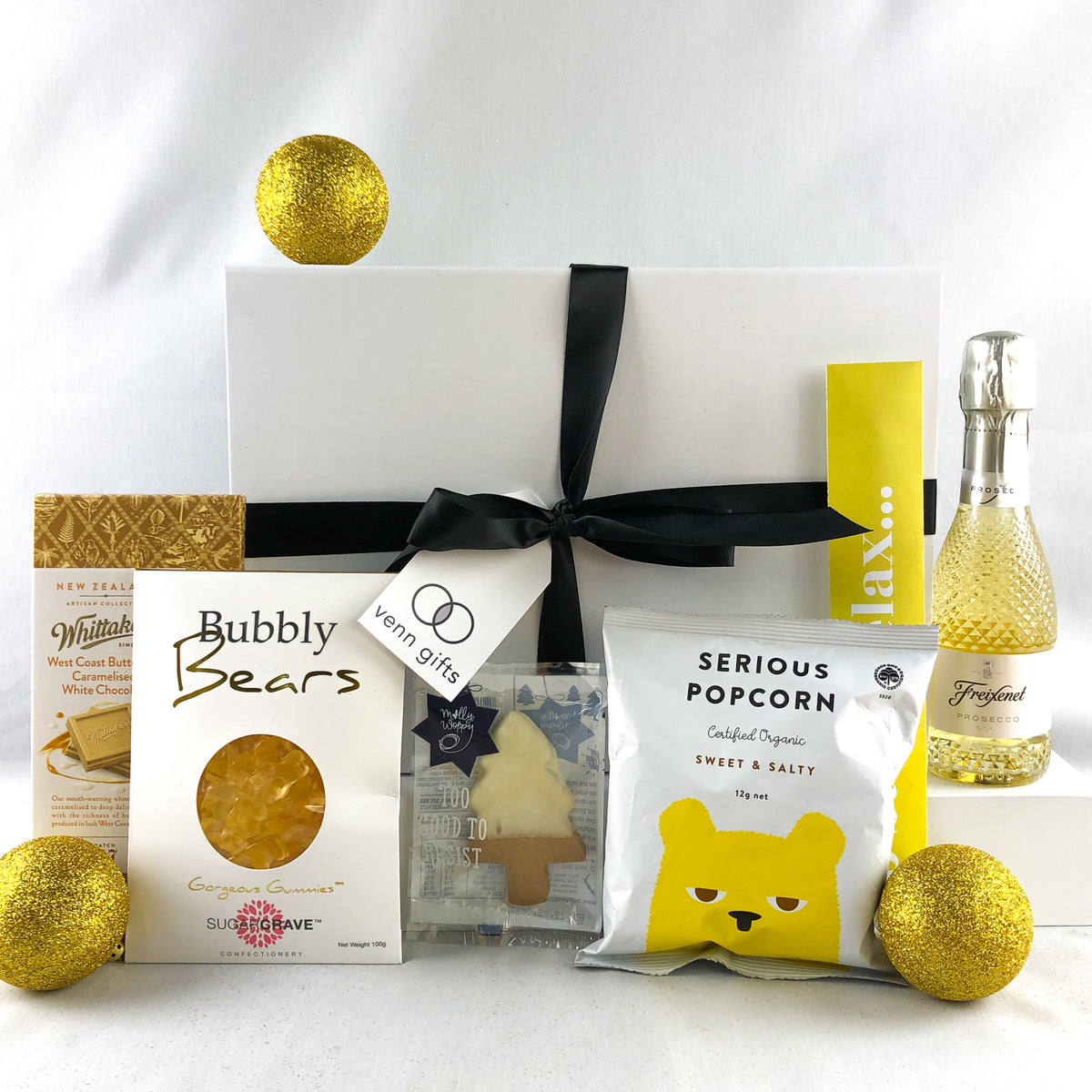 A curated gift box arrangement featuring a white box tied with a black ribbon, surrounded by assorted packaged snacks and treats including Bubbly Bears, Serious Popcorn, a bottle of Fréixenet sparkling wine, and gold glitter ornaments on a white background.