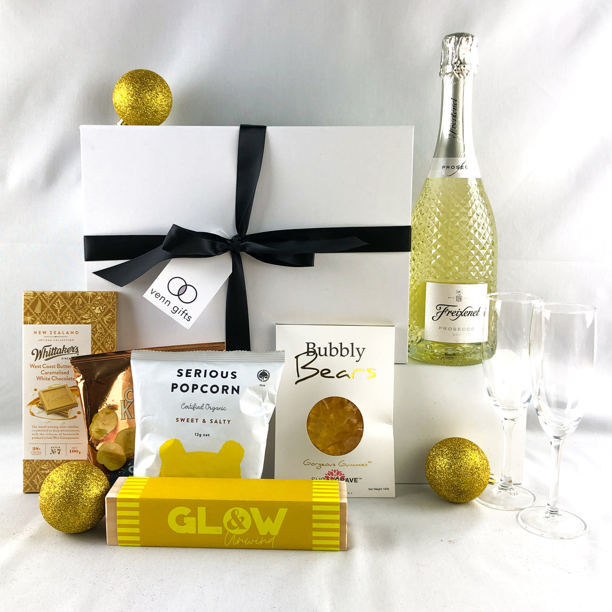 Freixenet prosecco, whittakers chocolate, popcorn, lollies and candles with glass flutes in front of a white giftbox with black ribbon against a white background by Venn Gifts.