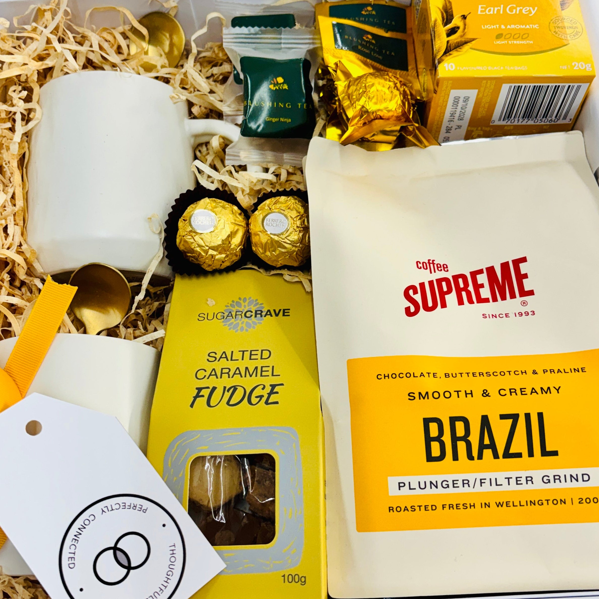 A curated gift box filled with assorted items on a white background. The box contains a white ceramic mug, a bag of Coffee Supreme Brazil coffee, a package of Salted Caramel Fudge, Twinings Earl Grey tea, gold foil wrapped chocolates, small jars, and shredded paper filler with a white gift tag.