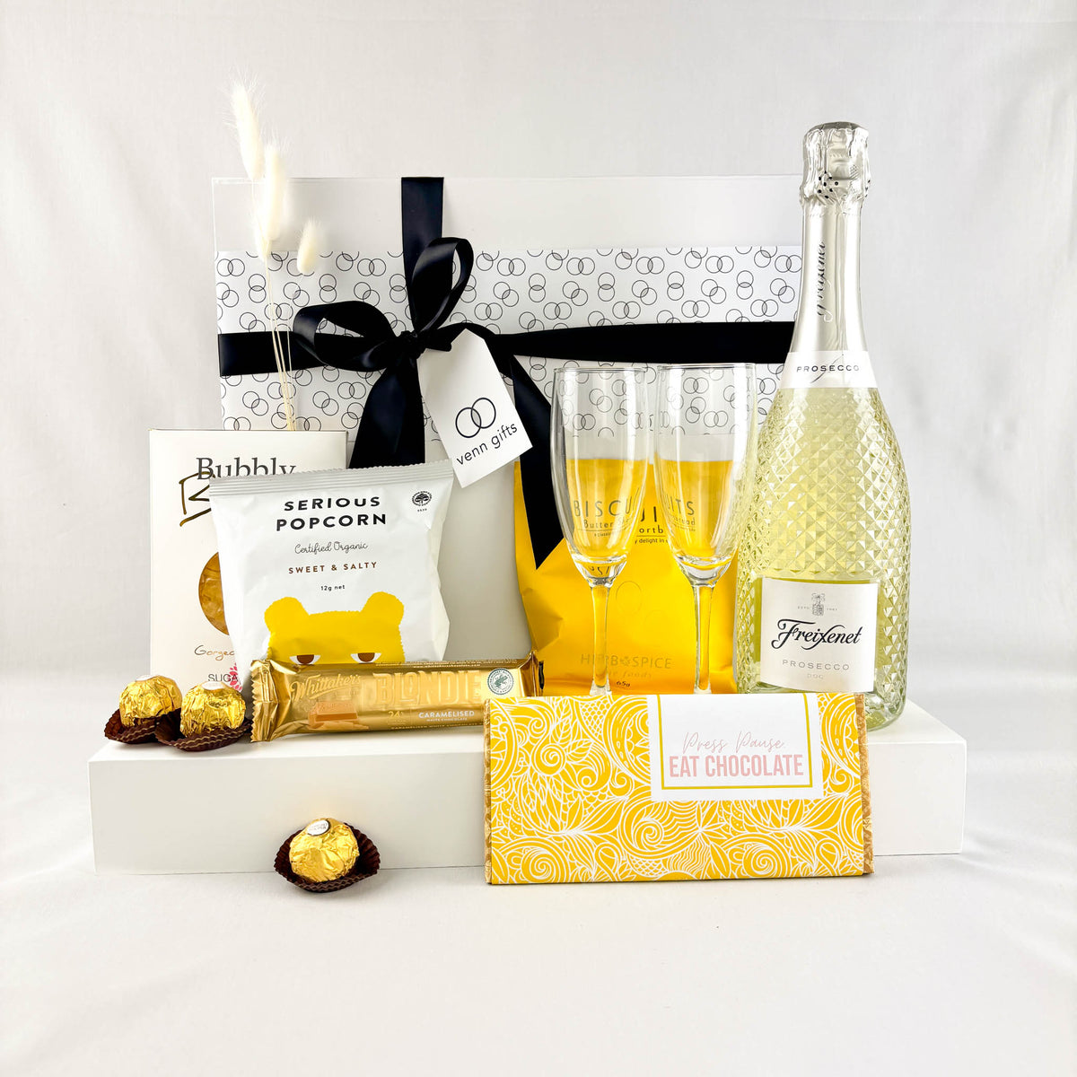 Freixenet 750ml bottle with two flutes, snacks and chocolates against a white gift box by Venn Gifts.