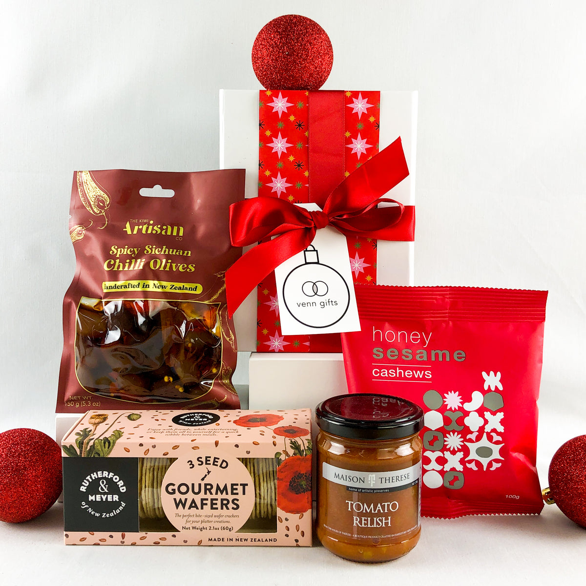 Red savoury snacks arranged around a white gift box with black ribbon by Venn Gifts.