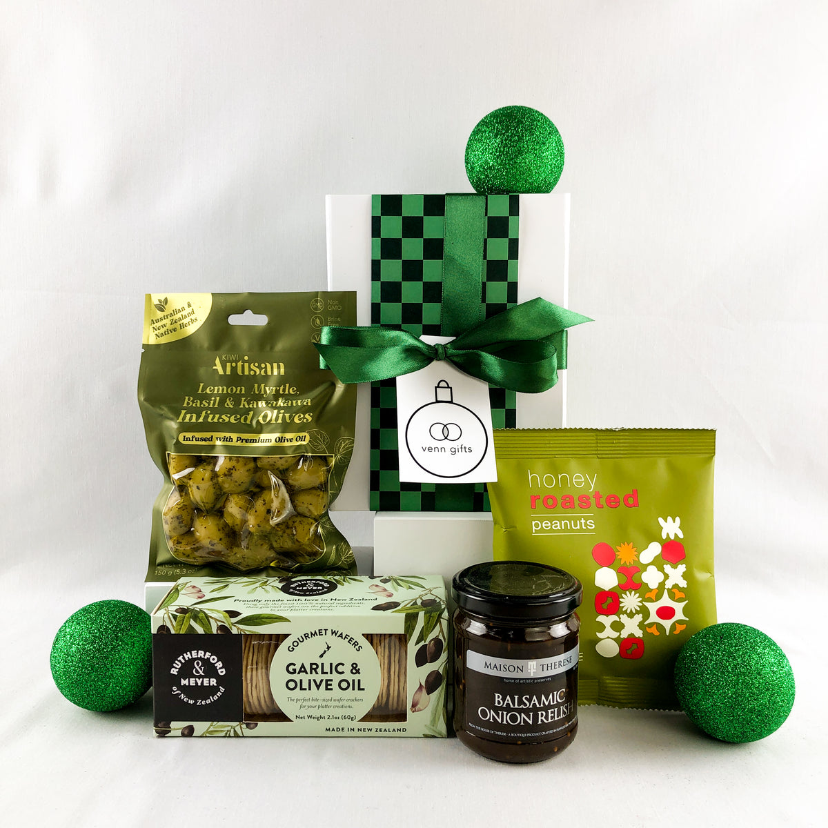 A curated gift box featuring gourmet snacks and treats, including a green and black checkered box with a green ribbon, artisan lemon olive oil and olives, honey roasted peanuts, and a jar of balsamic onion relish, styled with green decorative balls on a white background by venn gifts.