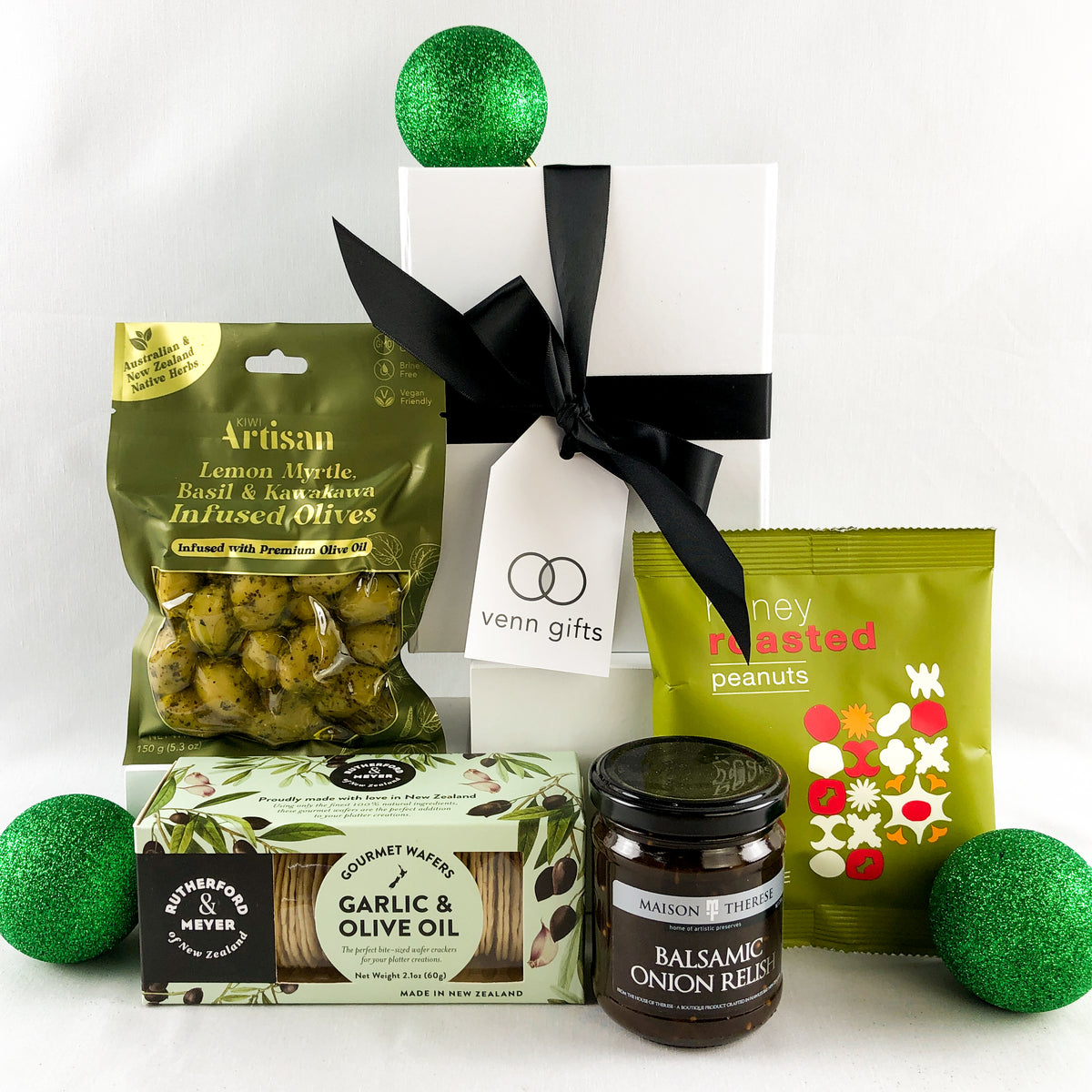 A curated gift box featuring gourmet snacks and treats including artisan lemon olive oil and olives, honey roasted peanuts, and a jar of balsamic onion relish, styled with green decorative balls on a white background by Venn Gifts.