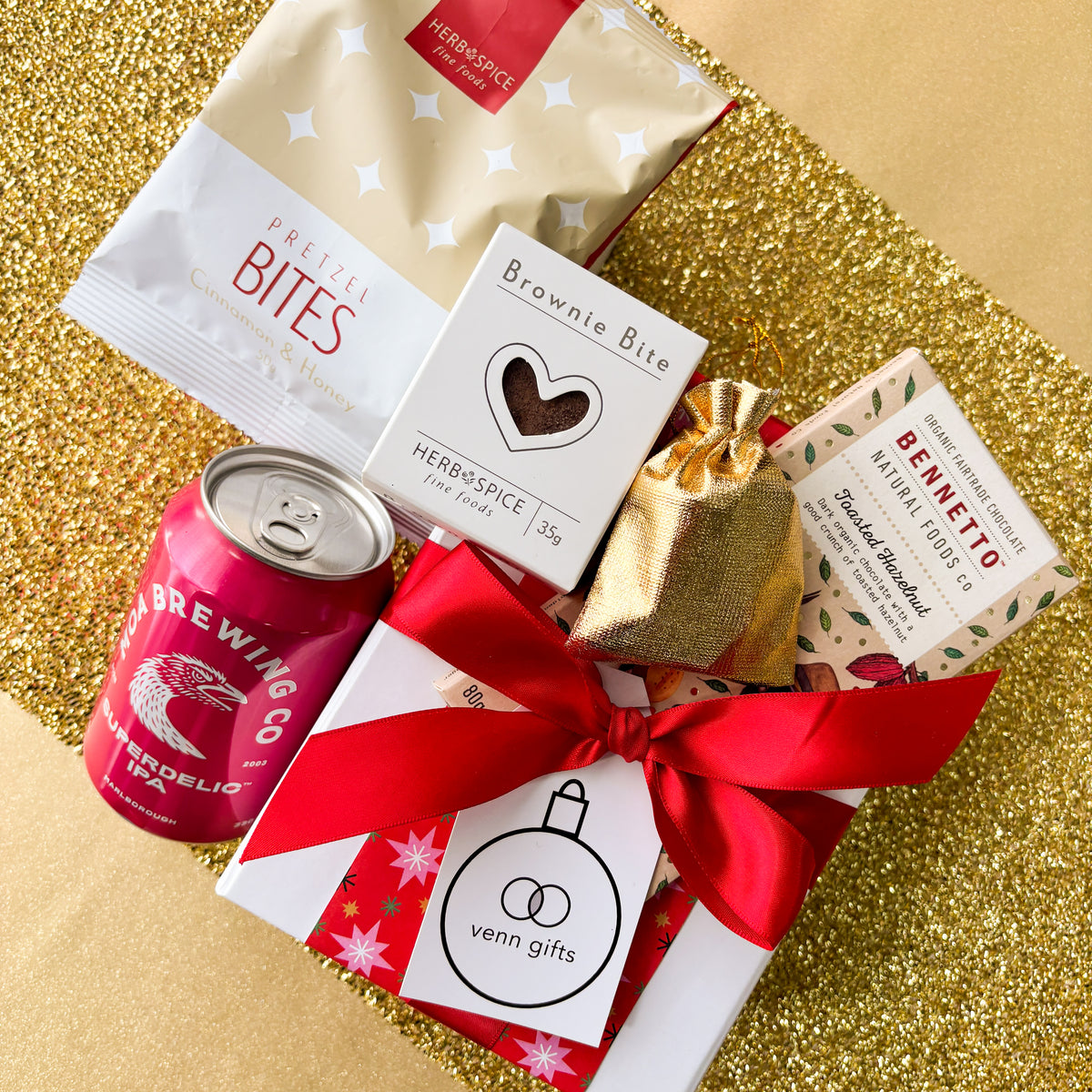 White giftbox overhead against a gold background. Containing pretzel bite, bennetto chocolatem moa brewing ipa, brownie bite, gold drawstring pouch filled with lollies from Venn Gifts.