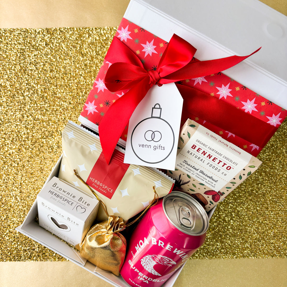 White giftbox overhead against a gold background. Containing pretzel bite, bennetto chocolatem moa brewing ipa, brownie bite, gold drawstring pouch filled with lollies from Venn Gifts.