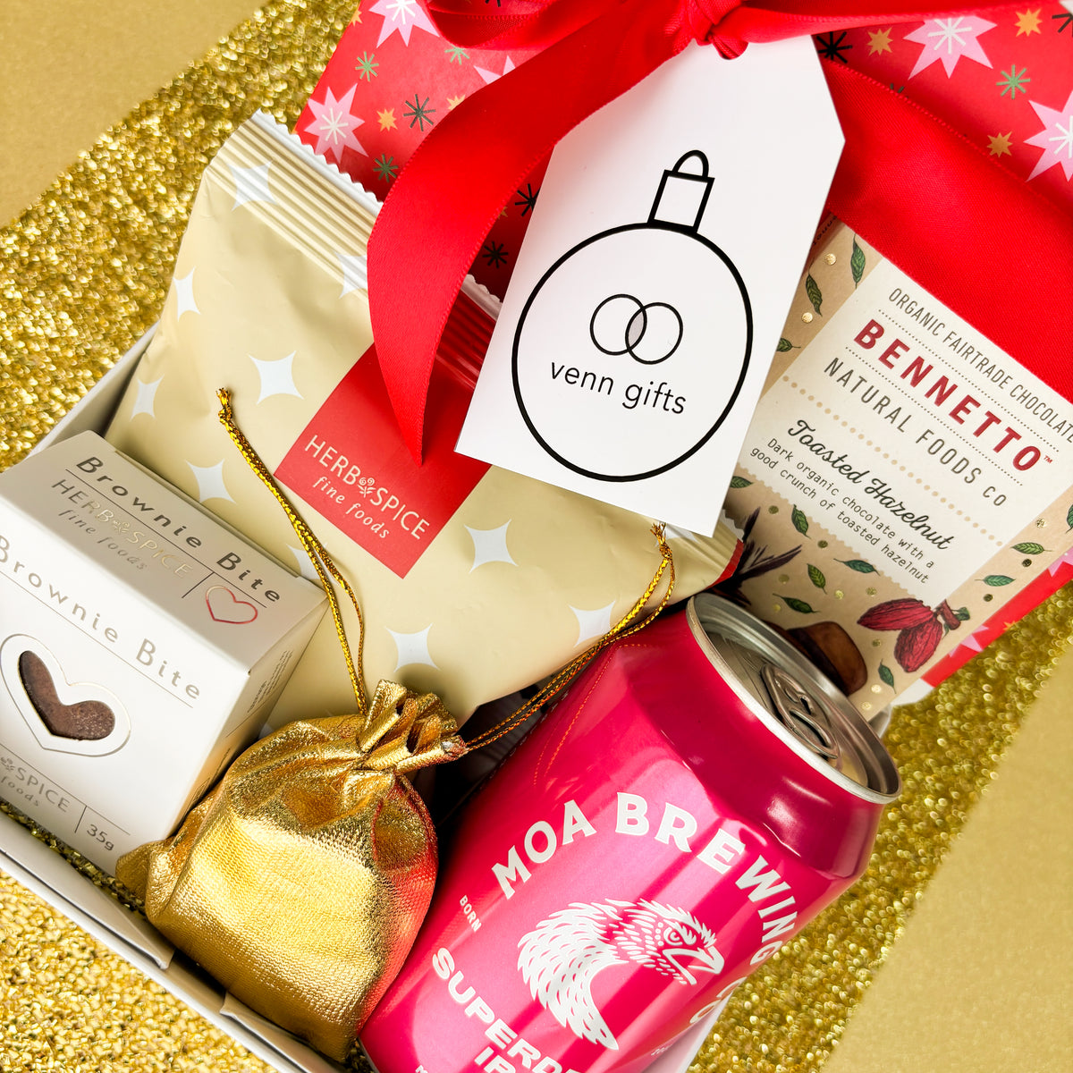 White giftbox overhead against a gold background. Containing pretzel bite, bennetto chocolatem moa brewing ipa, brownie bite, gold drawstring pouch filled with lollies from Venn Gifts.