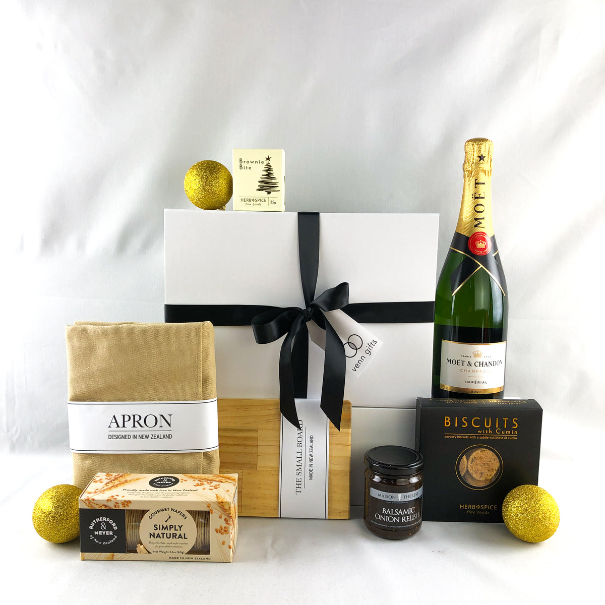 moet champagne with biscuits, crackers and relish alongside an apron and hopping board and christmas brownie against a white gift box with black ribbon and plain background by Venn Gifts.