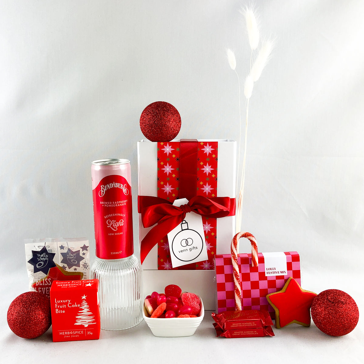 red drinks and snacks arranged around a white gift box with red ribbon against a white background. Christmas themed by Venn Gifts.