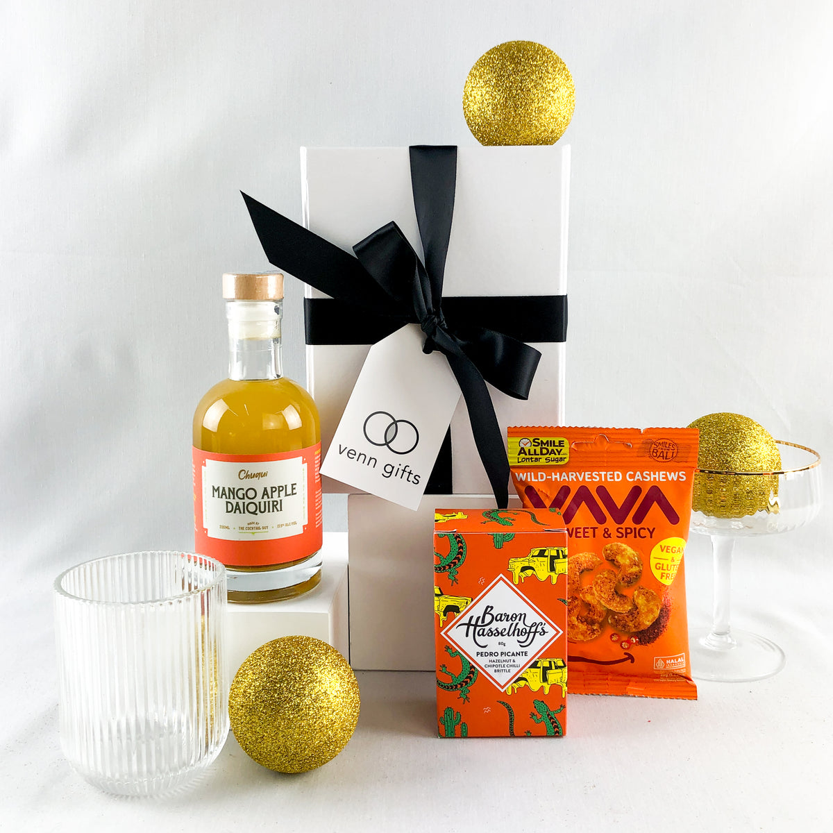 Daquiri, glass, nuts and baron hasselhoff's brittle displayed around a white giftbox with black ribbon against a white background by Venn Gifts