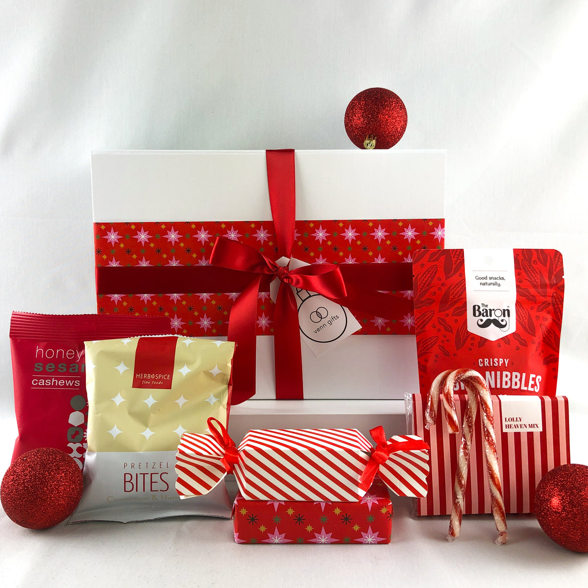A Christmas-themed gift box featuring a white box wrapped with a red ribbon and bow, surrounded by festive treats including red and white striped crackers, candy canes, and packaged snacks on a white background by Venn Gifts.