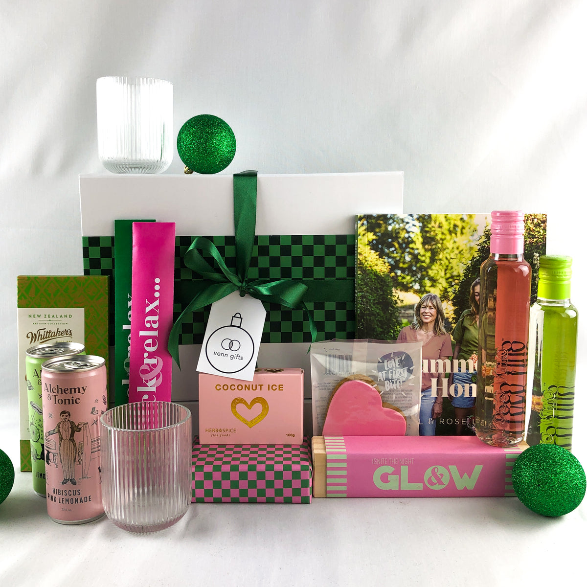 A curated gift box arrangement on a white surface with a white backdrop. Annabel Lanbein book and all other gifts are based around a green and pink colour theme in front of a white giftbox with green ribbon by Venn Gifts.