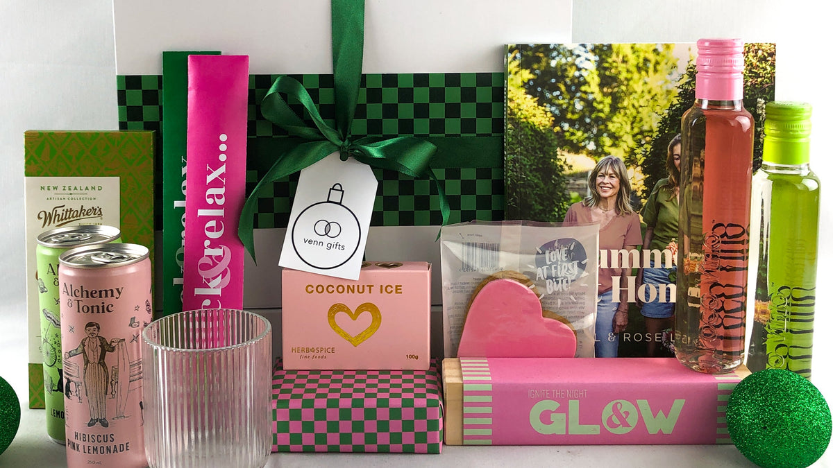 A curated gift box arrangement on a white surface with a white backdrop. Annabel Lanbein book and all other gifts are based around a green and pink colour theme in front of a white giftbox with green ribbon by Venn Gifts.