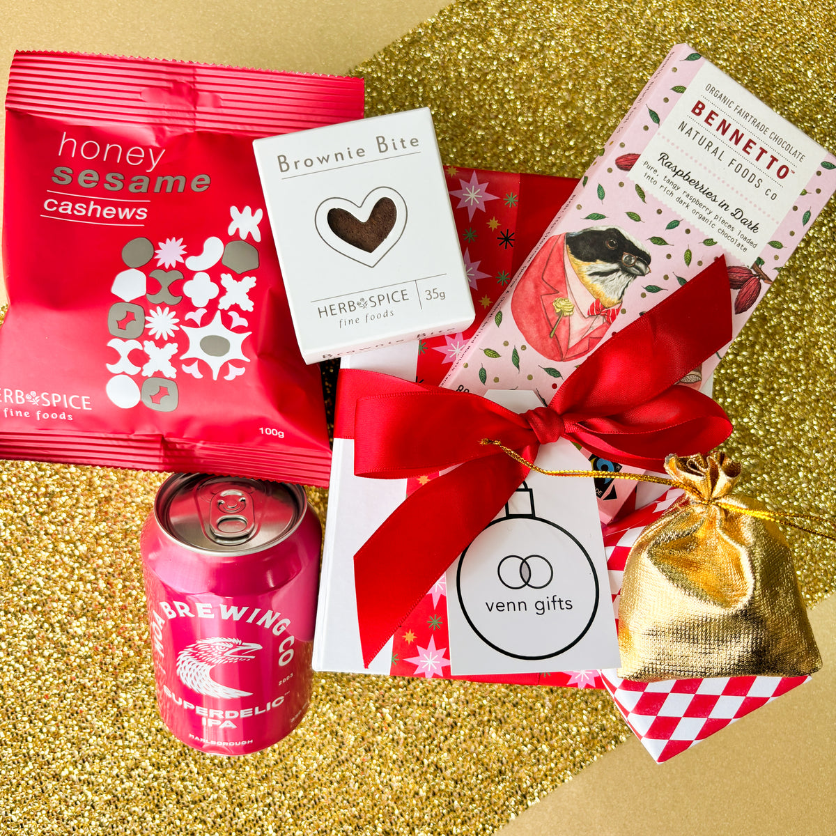 An assortment of chocolate, snacks and Moa Brewing IPA beer on a white giftbox with red ribbon against a gold background by Venn Gifts.