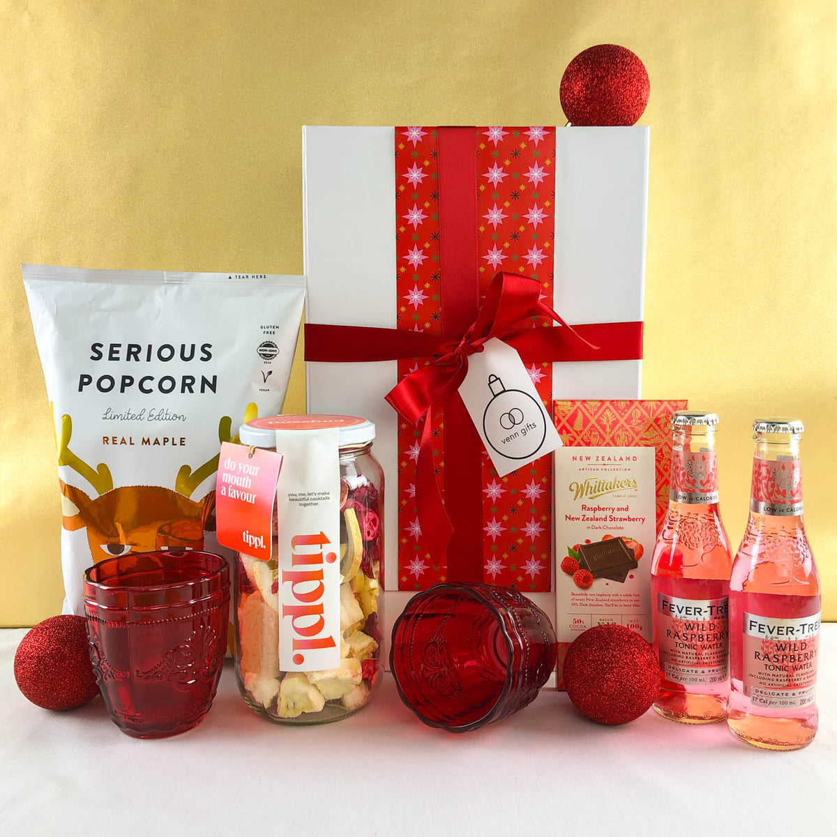 Tippl, Serious popcorn, Fever Tree & Whittaker's chocolate and red glasses surround a white gift box with a red bow against a gold background by Venn Gifts.