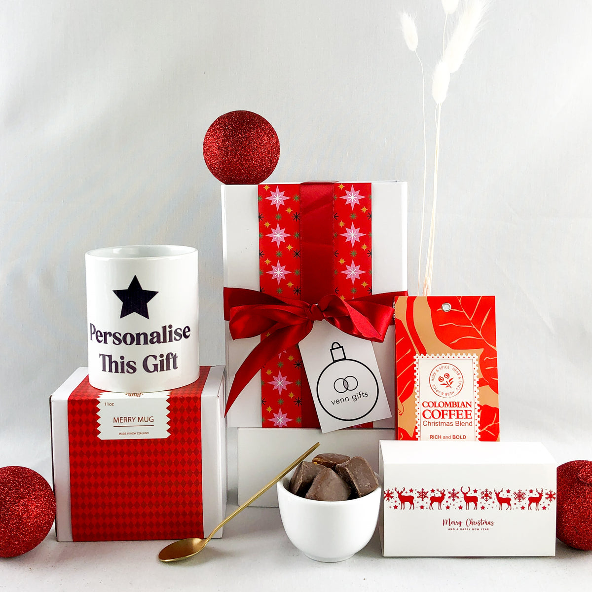 White giftbox, with christmas wrapping and red bow surrounded by a mug saying 'personalisse this gift', coffee and milk caramels with a gold teaspoon from Venn Gifts.