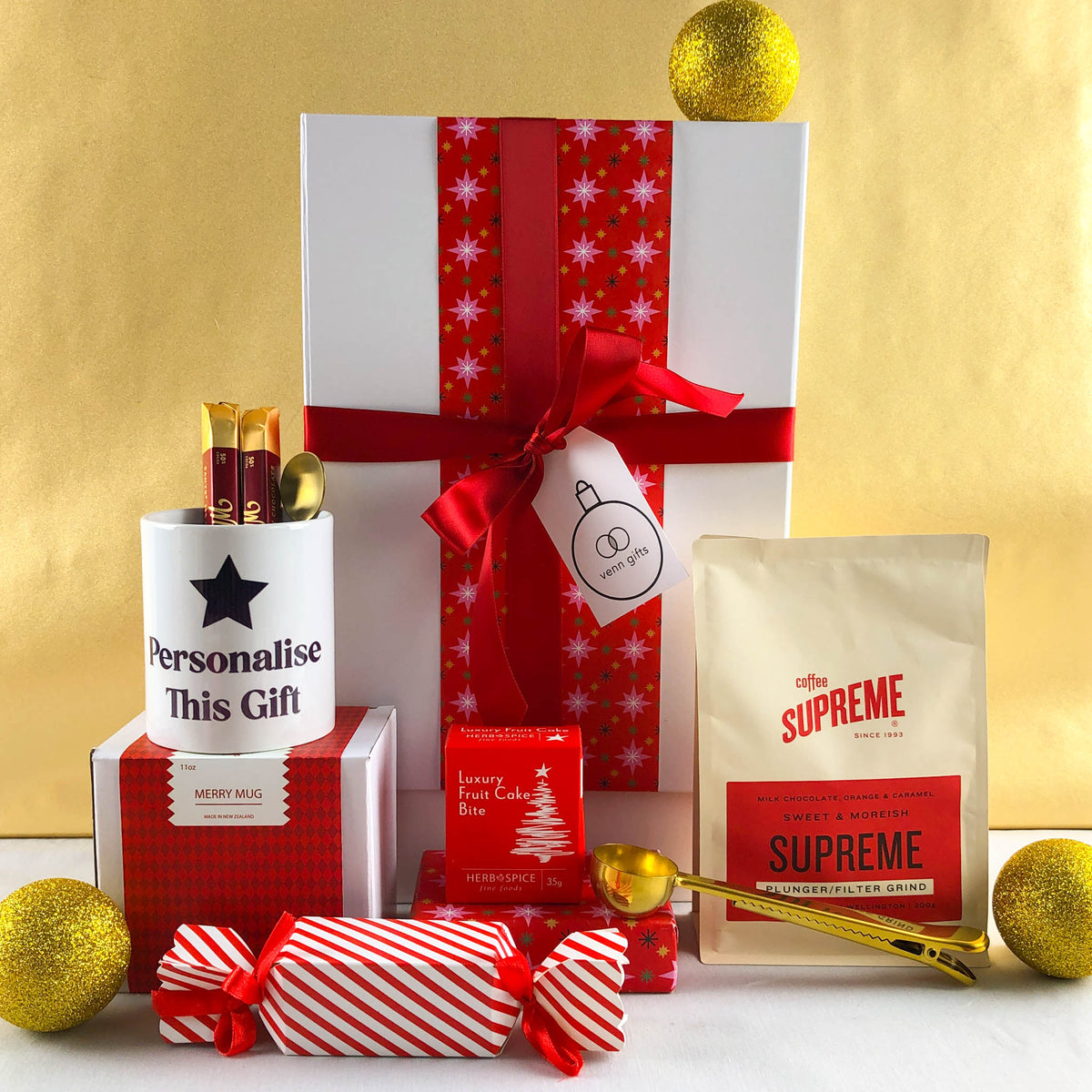 Personalised mug, coffee, Christmas cake, chocolates and fudge cake wrapped in red festive paper, surround a white gift box with a red ribbon against a gold background.