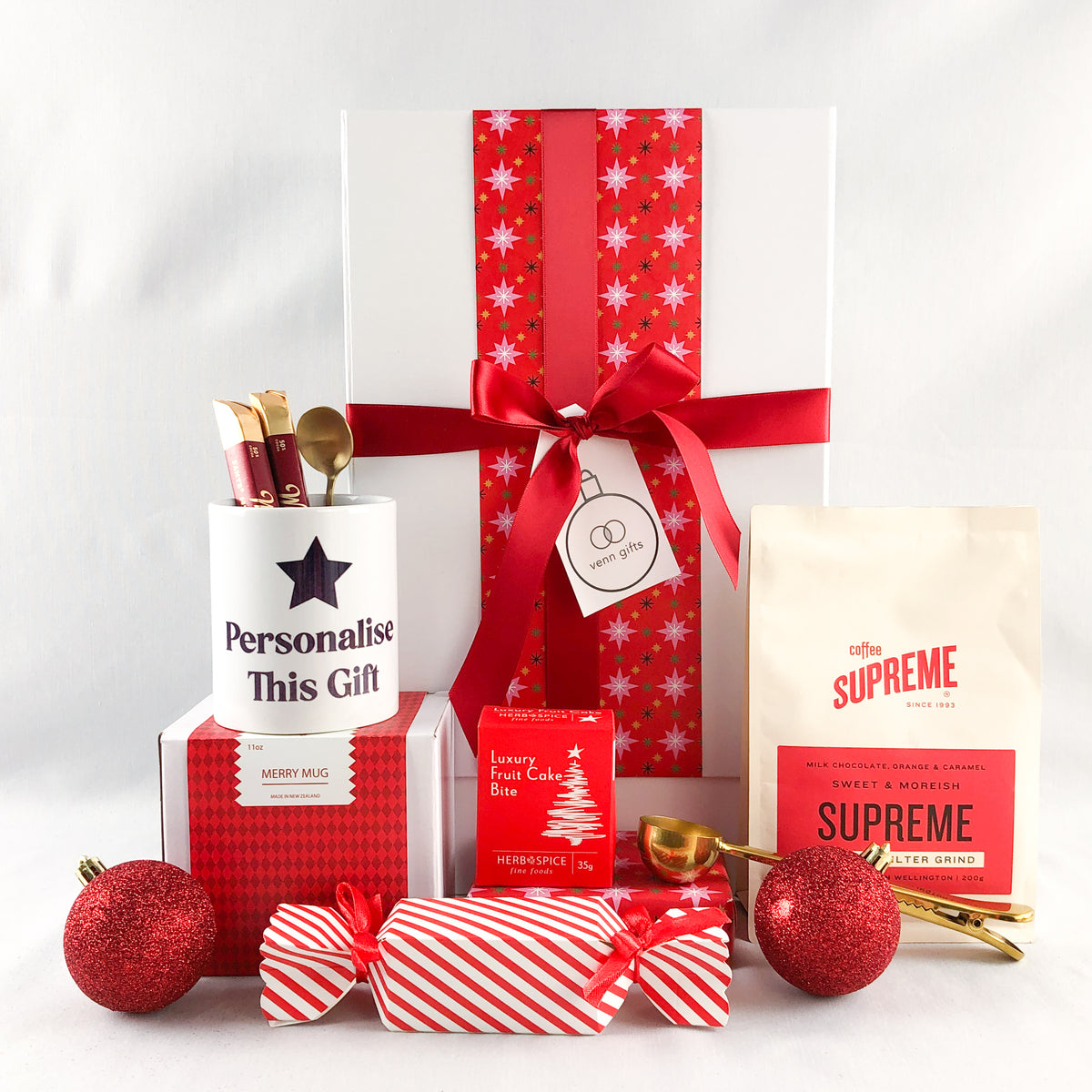 A Christmas-themed gift box arranged on a white background, featuring a large white gift box wrapped in red snowflake-patterned paper with a red ribbon and bow, a bag of Supreme coffee, a red patterned box, a white mug printed with 'Personalise This Gift', wrapped candy canes , gold spoon, and christmas cake and cracker by Venn Gifts.