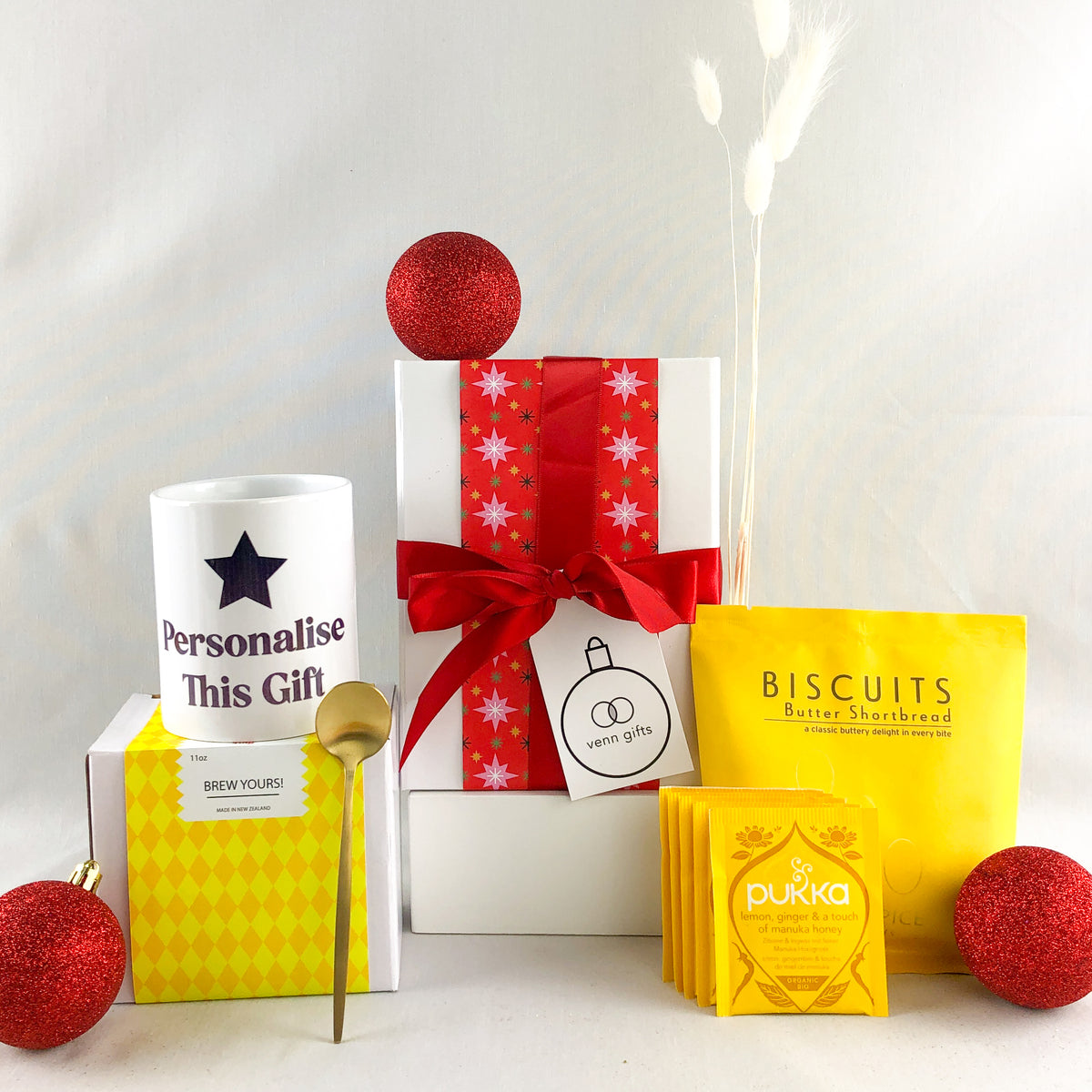 Personalised mug with teaspoon, yellow packaged butter shortbread and pukka teas set around a white giftbox with red ribbon by Venn Gifts.
