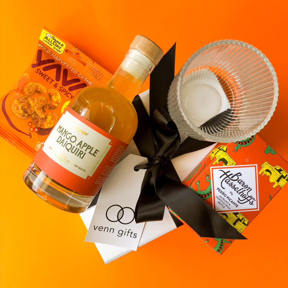 Daquiri, glass, nuts and baron hasselhoff's brittle on top of a white giftbox with black ribbon against an ornage background by Venn Gifts