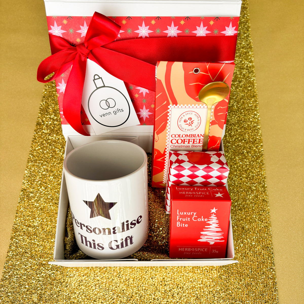 White gift box with red wrapping and red bow with coffee, gold teaspoon and personalised mug and gift wrapped christmas fruit cake against a gold background by Venn Gifts.