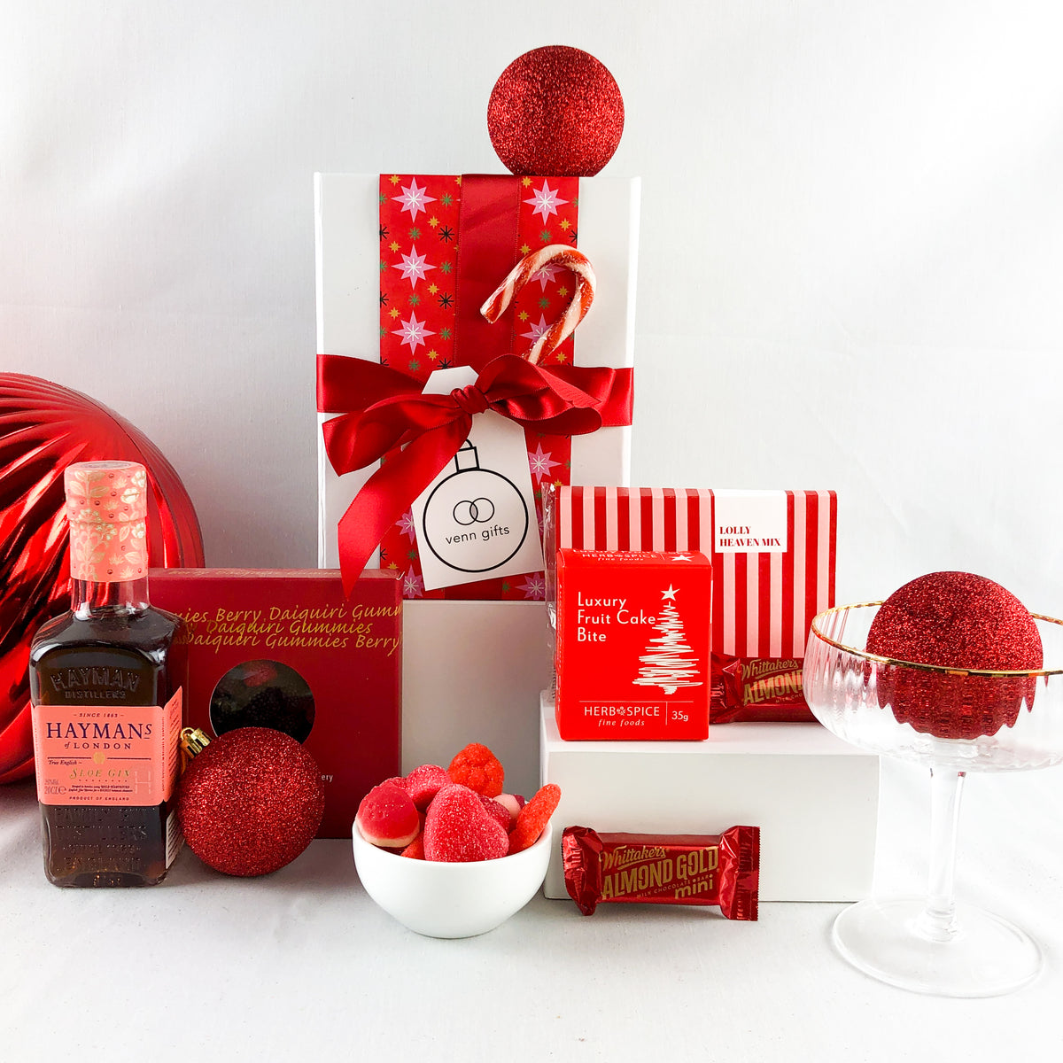 A curated Christmas gift box arrangement on a white surface against a white background, featuring a bottle of Haymans London gin, red and white striped gift packaging, a red chrsitmas ornament, a bowl of strawberry lollies and Whittaker's chocolates by Venn Gifts.