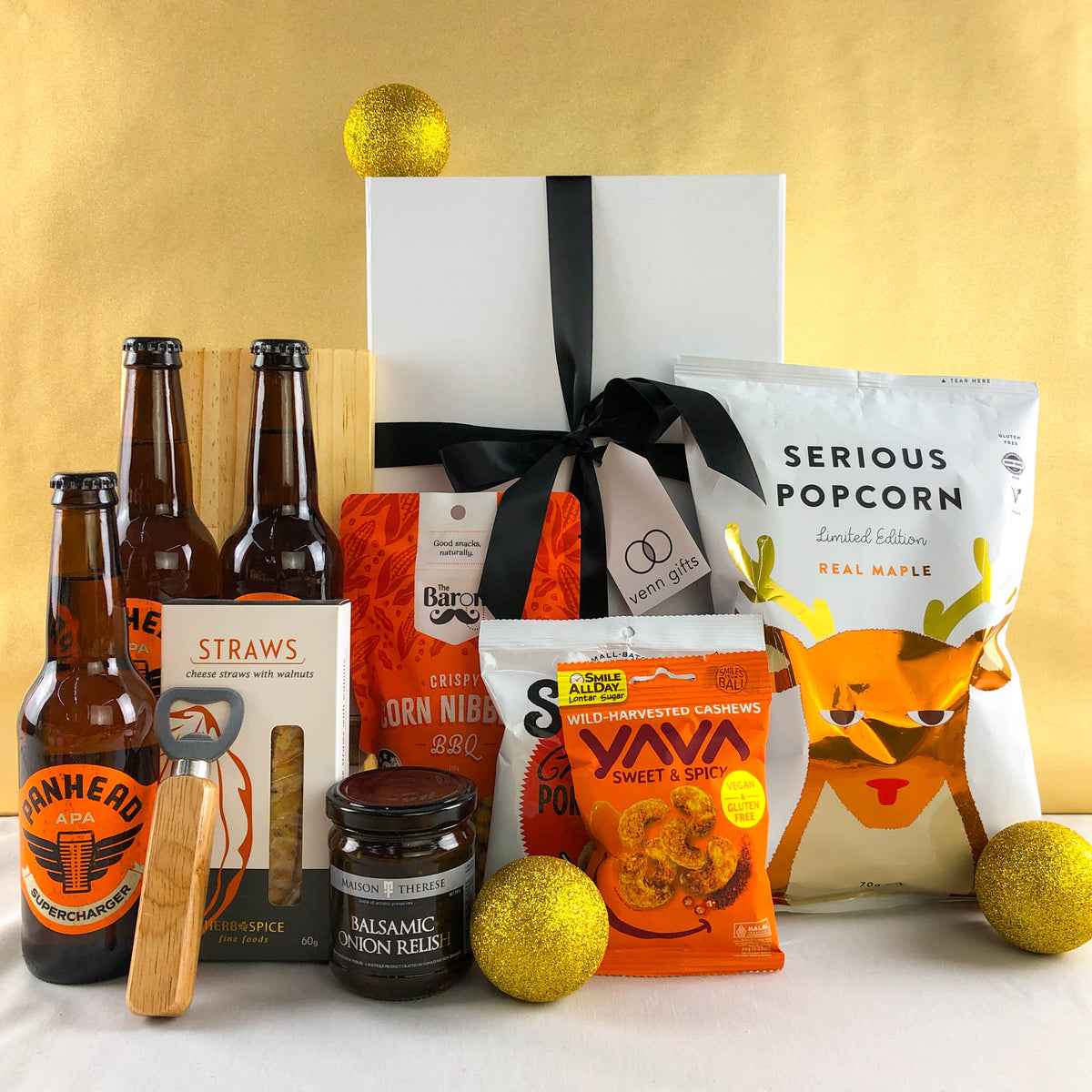 Beers, popcorn, snacks surround a white gift box with black bow against a gold ribbon.