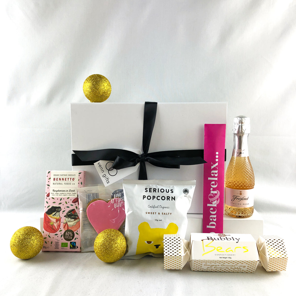 Snacks, treats including Bennetto chocolate, serious popcorn and lollies and Freixenet rose wine  in front of a white gift box with black ribbon against a white background by Venn GIfts.