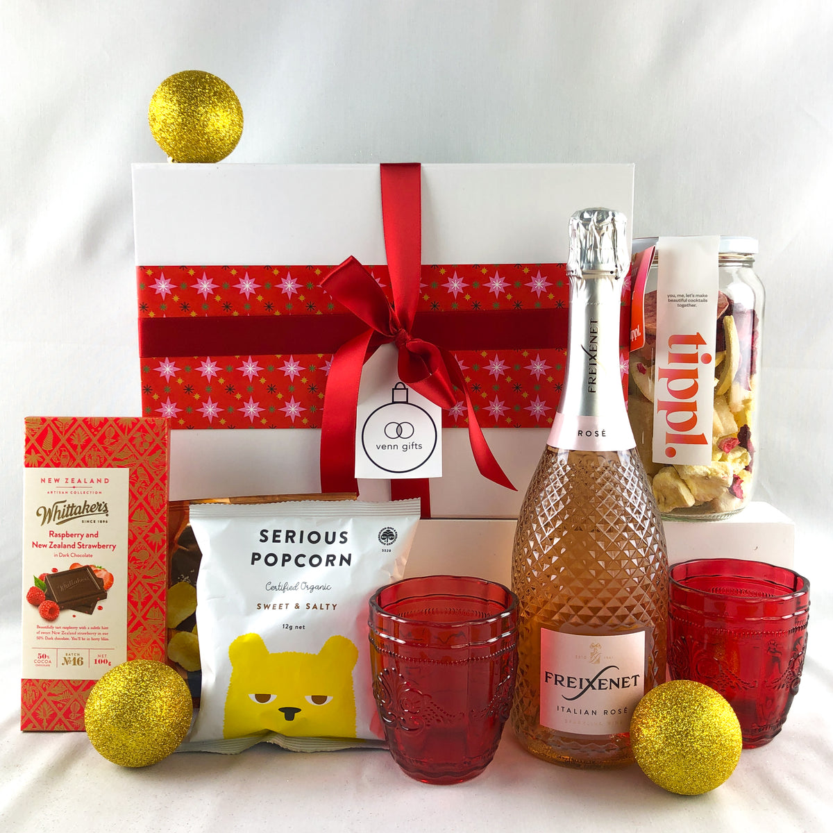 A curated gift box arrangement on a white fabric background, featuring a white gift box with red snowflake pattern and ribbon, a bottle of Freixenet Rosé sparkling wine, two red textured drinking glasses, gold glitter ornaments, a bag of Serious Popcorn, Tippl cocktail infusion by Venn Gifts.