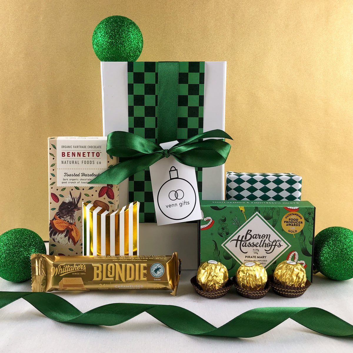 White gift box with green ribbon surrounded by chocolates by Baron Hasselhoff, Bennetto, Whittaker's, Ferrerro Rocher & Chocolate Brownie against a gold background by Venn Gifts.