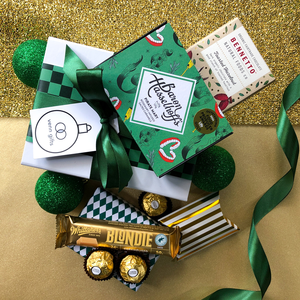 White gift box with green ribbon surrounded by chocolates by Baron Hasselhoff, Bennetto, Whittaker's, Ferrerro Rocher & Chocolate Brownie against a gold background by Venn Gifts.