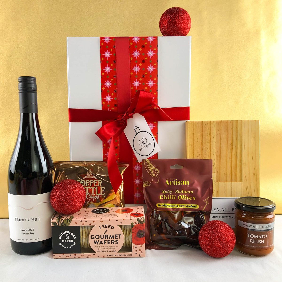 Trinity Hill red wine, olives, Maison Terese chutney and other snacks and chopping board surround a white gift box with red ribbon against a gold backgound by Venn gifts.