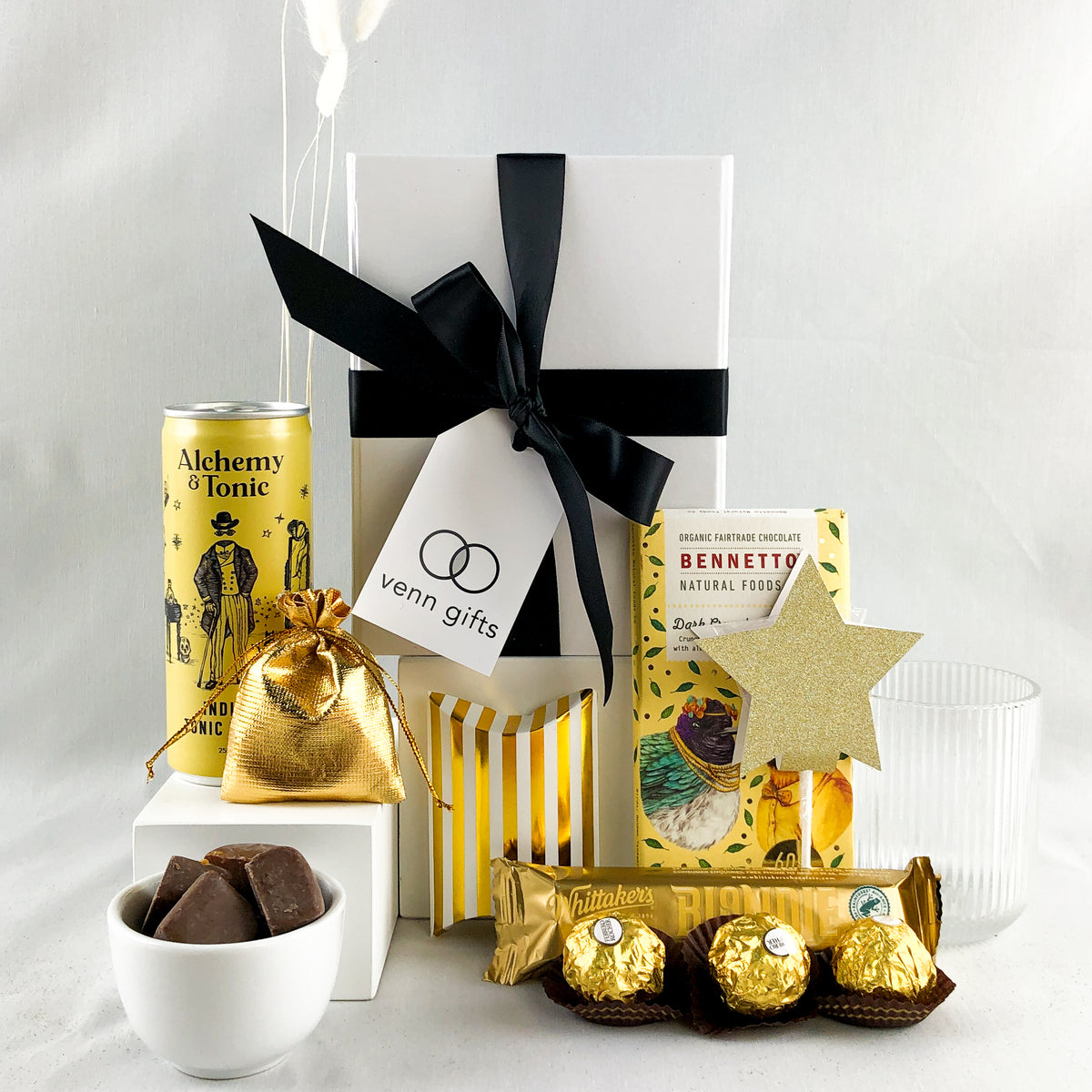 A curated gift box displaying a drink and glass, snacks and chocolates including Bennetto, Ferrerro Rocher & Whittakers all in gold or yellow colour ways. Presented in a white gift box with black ribbon by Venn Gifts.