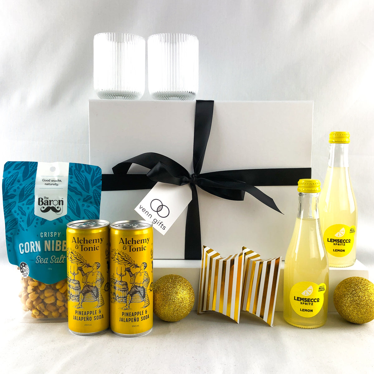 A curated gift box set on a white background, featuring a white gift box tied with a black ribbon, two white ribbed glasses a bag of Baron Crispy Corn Nibs, two Alchemy & Tonic can drinks, two lemonsecco bottles by Venn Gifts.