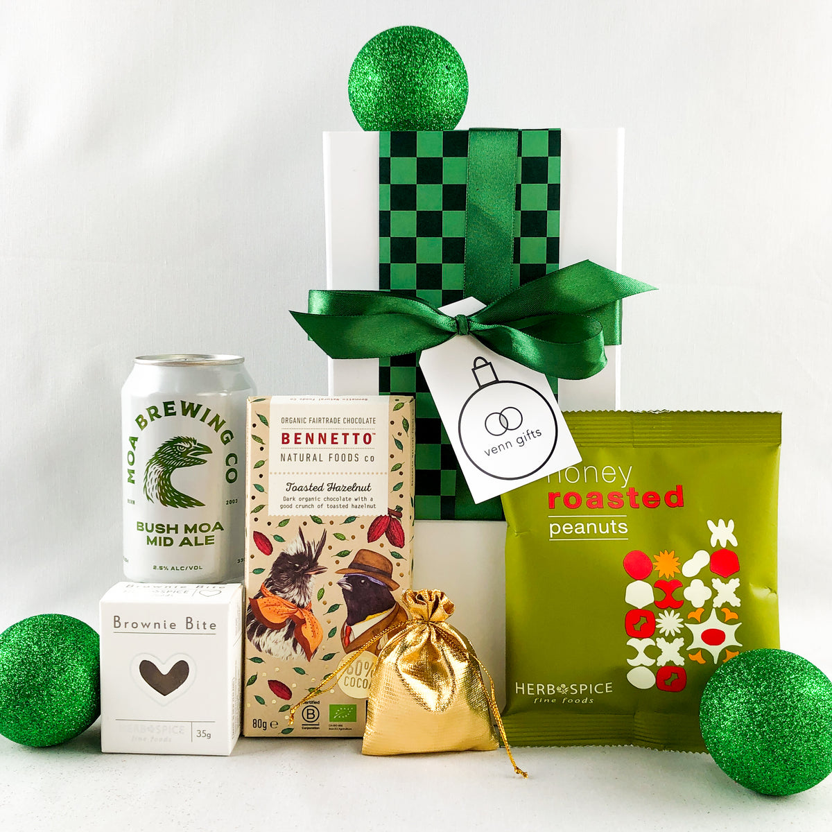 A curated gift  arrangement on a white background featuring a white gift box wrapped in green checkered paper with a green ribbon and bow, a can of Moa Brewing Co. Bush Moa Mid Ale, a box of Bennetto chocolate, a green bag of  Honey Roasted Peanuts, a small gold drawstring pouch, and green glitter christms decorations by Venn Gifts.
