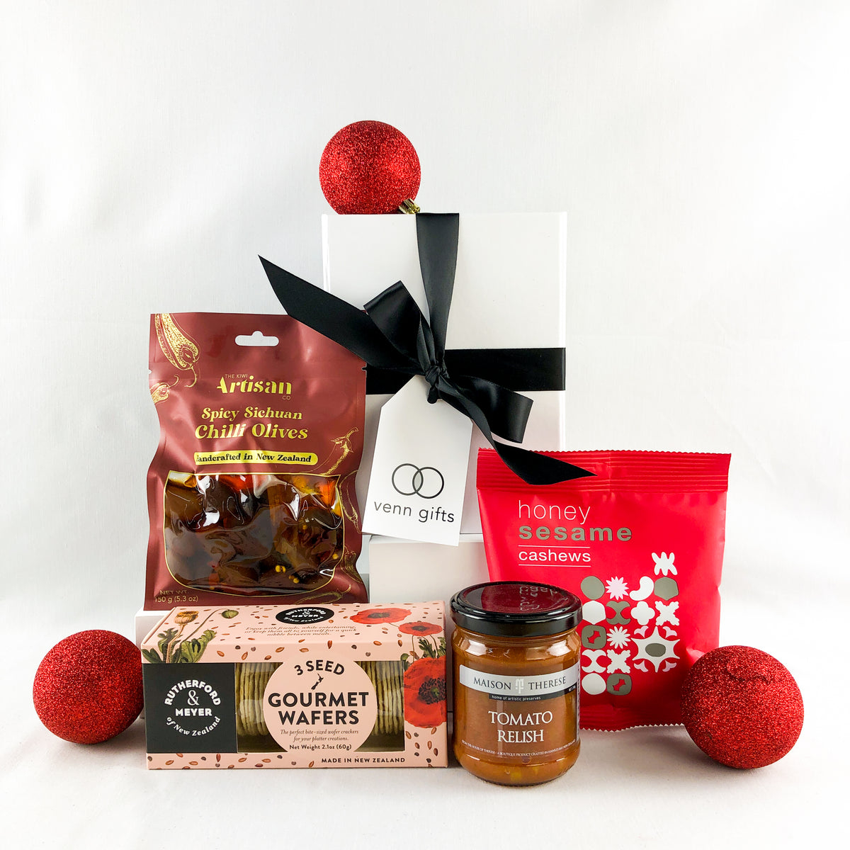 Red savoury snacks arranged around a white gift box with black ribbon by Venn Gifts.