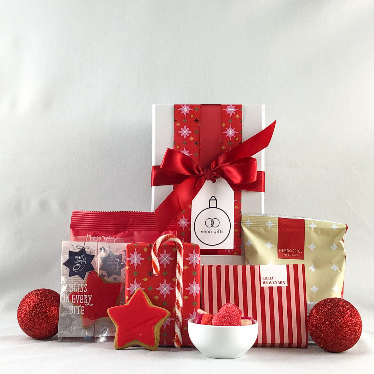A Christmas gift box featuring red and white striped packaging, a large red ribbon bow, a star-shaped cookie, candy canes, red wrapped treats, and wrapped chcoolate fudge cake against a white gift box with red ribbon by Venn Gifts.