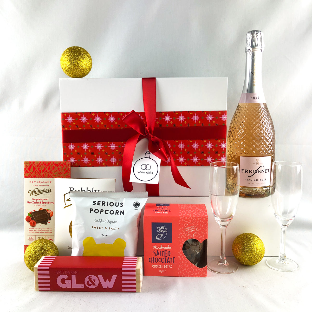 A festive gift hamper set on a white fabric background, featuring a large white gift box with a red ribbon and snowflake pattern, a bottle of Frexenet Rose Sparkling Wine, two champagne flutes, a bag of Serious Popcorn, a box of Salted Chocolate cookies, a bar of whittakers chocolate, lollies and a candle holder by Venn Gifts.