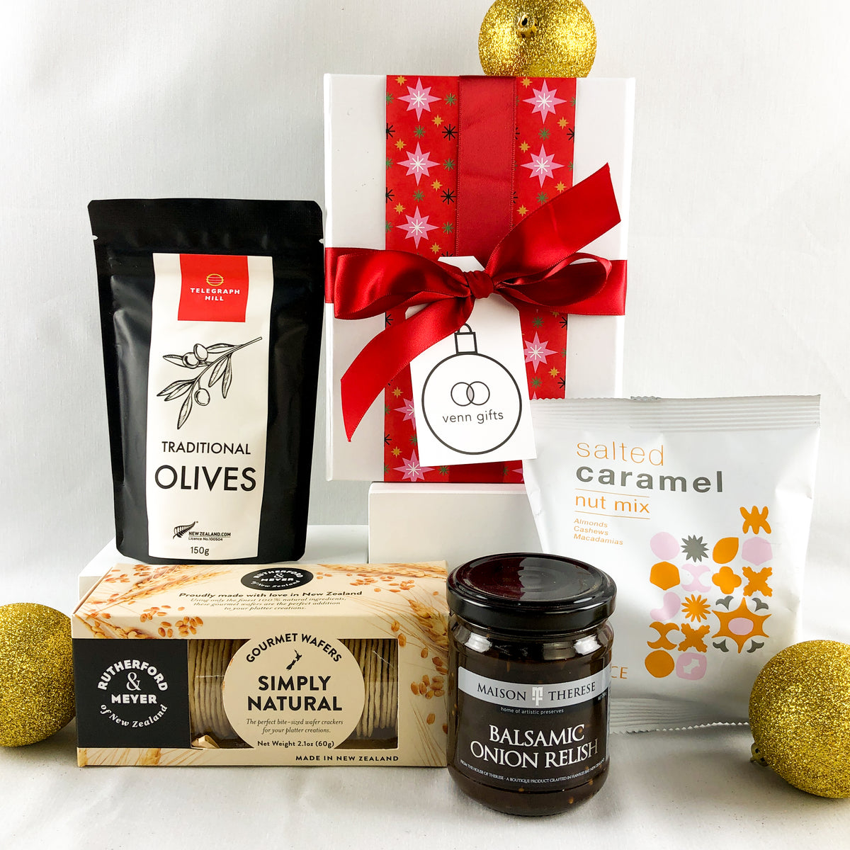 Olives, relish, nuts and crackers arranged in front of a white giftb ox with a red ribbon against a white background by Venn Gifts.
