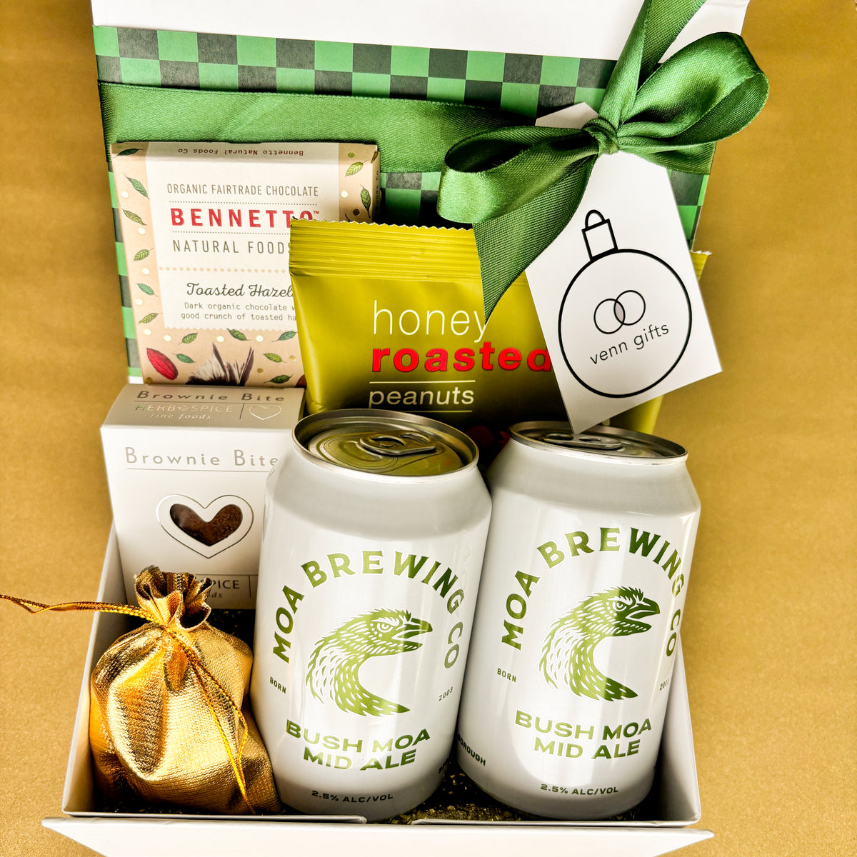 A curated gift box set containing 2 cans of Moa Brewing Co. Bush Moa Mid Ale, a box of Bennetto Chocolate, honey roasted peanuts, a brownie bite, and a small gold ribbon bag, arranged in a white and green checkered box with a green ribbon by Venn Gifts.