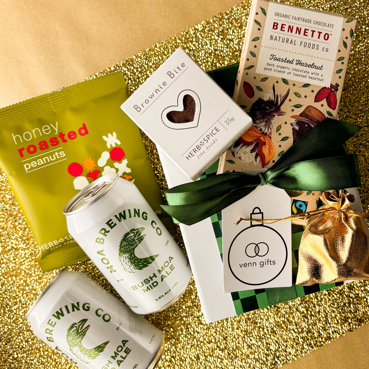 A curated gift basket on a gold glitter background, containing a green bag of Honey Roasted Peanuts, two white cans of Moa Brewing Co. beer, a white box of Brownie Bite, a Bennetto chocolate bar and a wrapped gift box tied with a dark green ribbon and a small gold drawstring pouch by Venn Gifts.