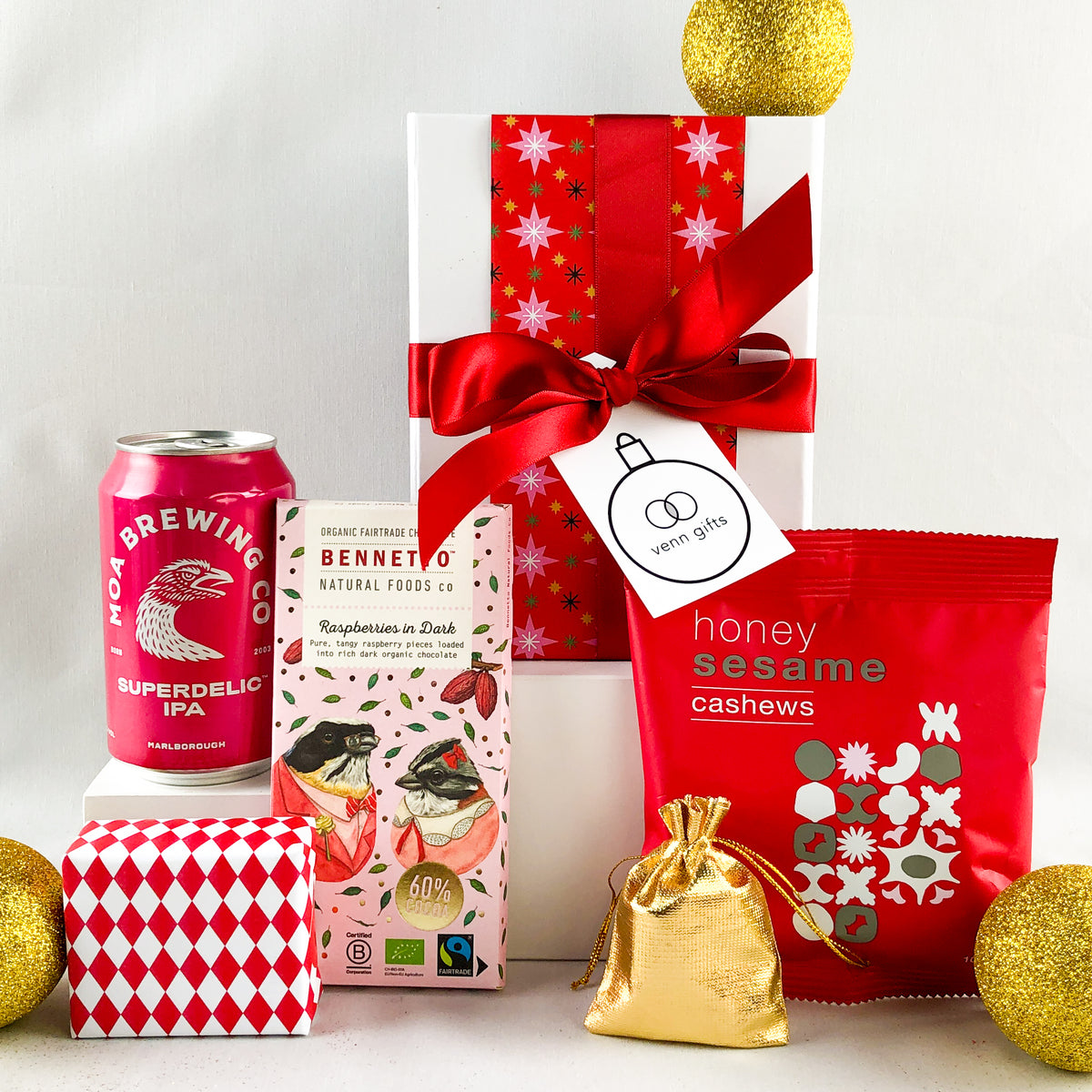 A curated gift  box wrapped in red patterned paper and tied with a large red ribbon, alongside a can of red moa brewing beer, Bennetto chocolate, a red honey sesame cashews pouch, a small gold drawstring pouch, and gold christmas decorations by Venn Gifts.