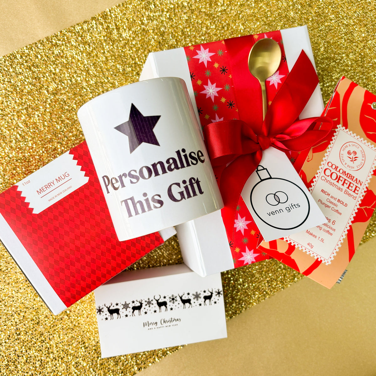White giftbox, with christmas wrapping and red bow surrounded by a mug saying 'personalise this gift', coffee and milk caramels with a gold teaspoon on a gold background from Venn Gifts.