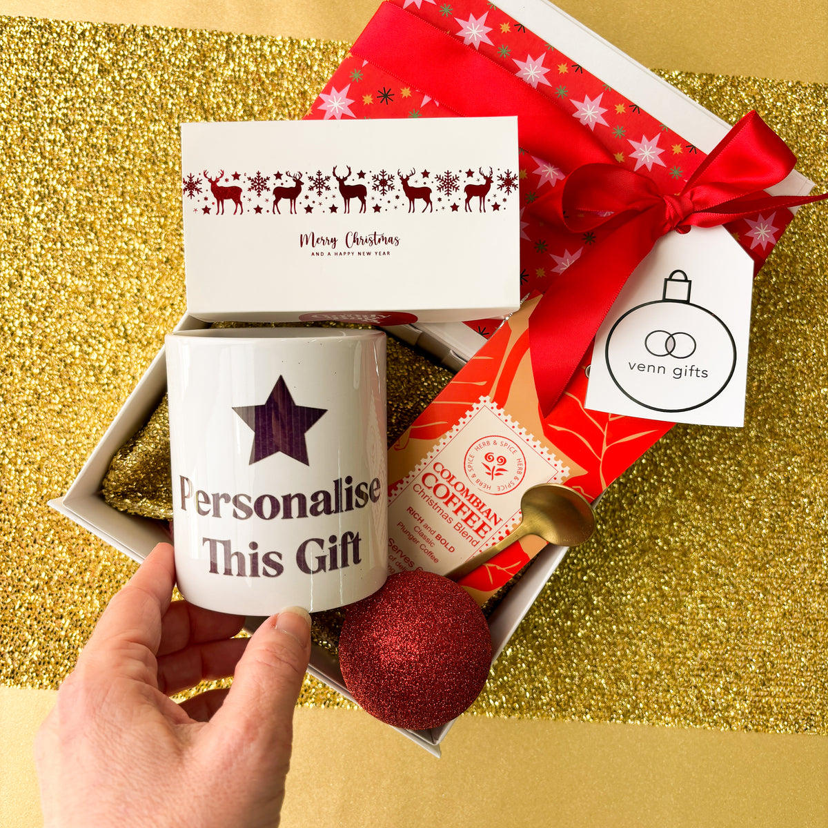 White giftbox, with christmas wrapping and red bow surrounded by a mug saying 'personalise this gift', coffee and milk caramels with a gold teaspoon from Venn Gifts.