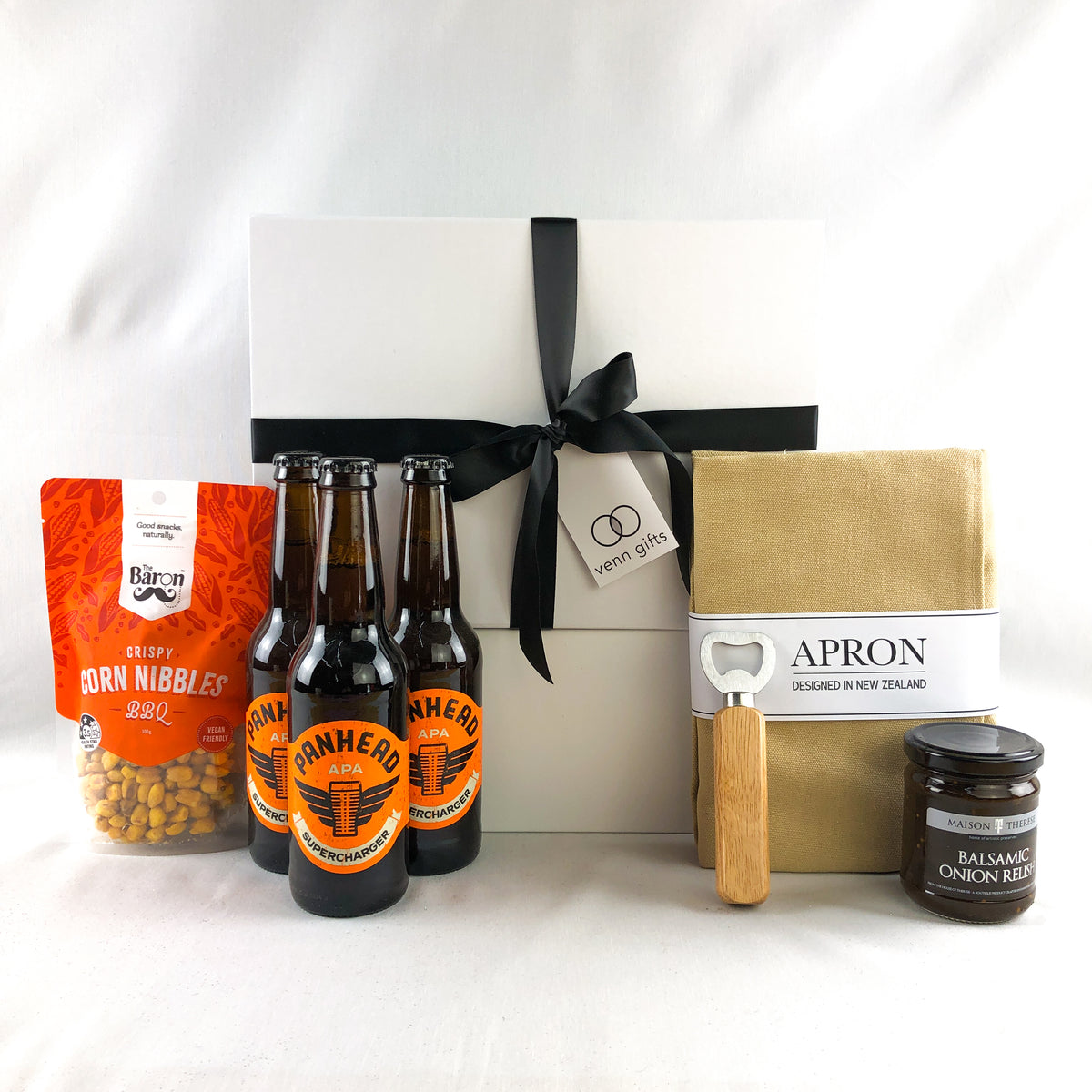 brown, orange and bronze colour gifts: apron, bottle opener, beers and BBQ corn nibbles with relish in front of a white giftbox with black ribbon by Venn Gifts
