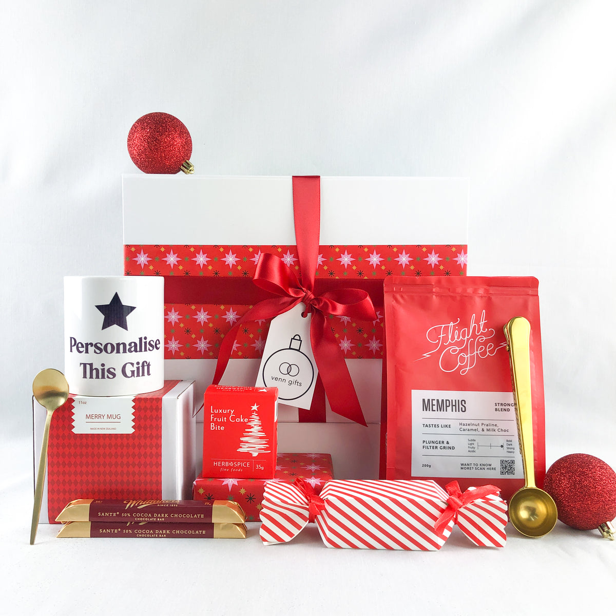 A Christmas-themed gift box arranged on a white background, featuring a large white gift box wrapped in red snowflake-patterned paper with a red ribbon and bow, a bag of Flight coffee, a red patterned box, a white mug printed with 'Personalise This Gift', wrapped candy canes , gold spoon, and christmas cake and cracker by Venn Gifts.