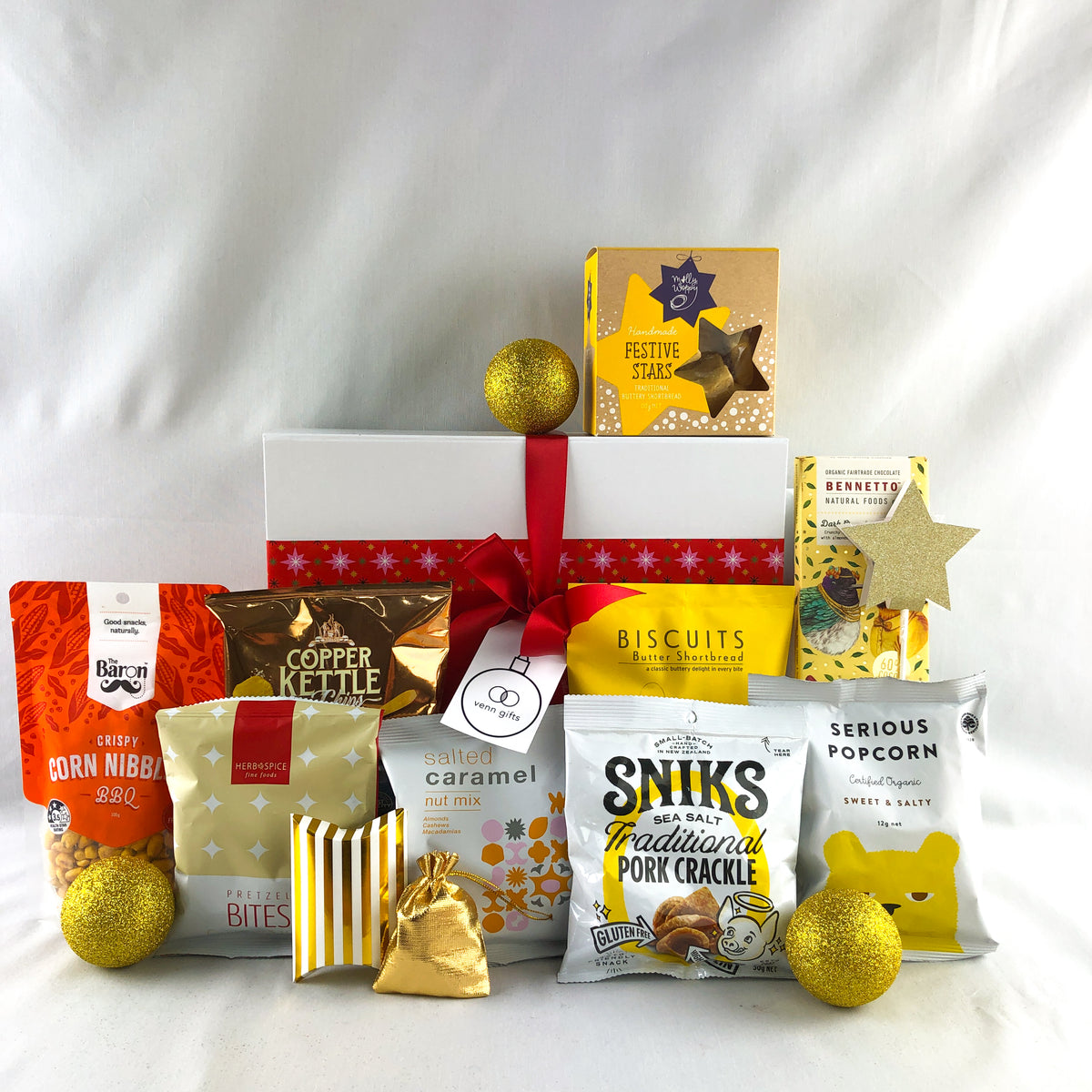 A festive gift box filled with assorted snack and treat packages, arranged in front of a white gift box tied with a red ribbon. Visible items include Snickers Caramel, Sniks Pork Cracker, Serious Popcorn, The Baron Corn Nibbles, Copper Kettle, and Festive Treats chocolate, with gold and red decorative accents in front of a white giftbox with red ribbon by Venn Gifts